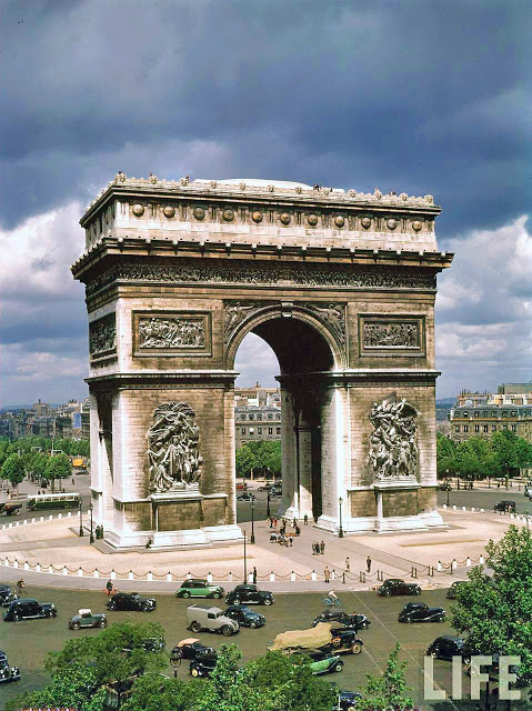 a large stone arch with statues and people around it with Arc de Triomphe in the background
