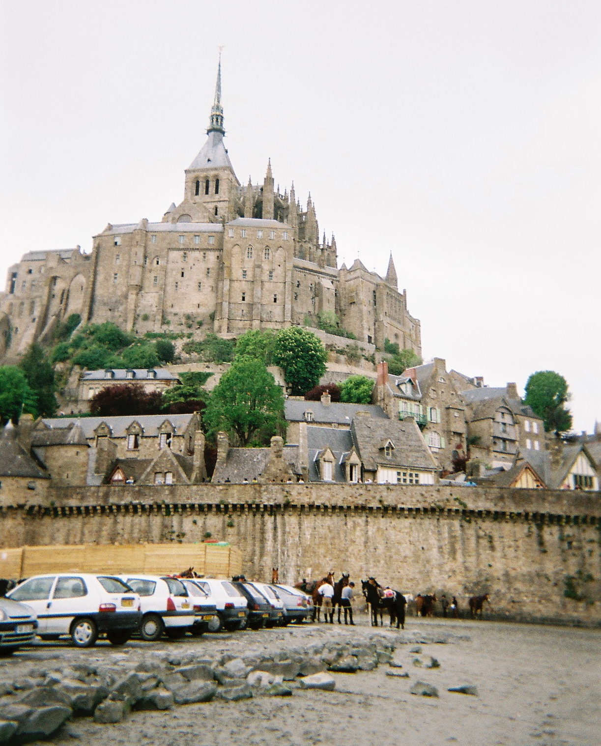 a castle on a hill with horses and cars parked in front of it with Mont Saint-Michel in the background