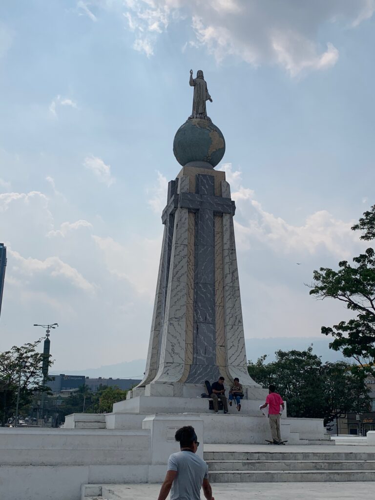 a statue of a man on a pedestal with Monumento al Divino Salvador del Mundo in the background